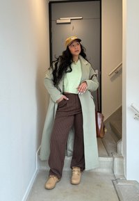 Lange lichtgroene jas over een muntgroene trui, bruine gestreepte broek en tan laarzen. Accessoires omvatten een tan pet en een bruine tas.