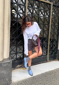 Mocassins azul claro com costura decorativa, combinados com meias cinzentas e uma saia riscada. O fundo apresenta uma porta preta texturizada.