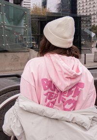 Roze hoodie met grote tekst op de achterkant, gelaagd onder een lichtgrijze jas. De persoon draagt een roomkleurige muts en de omgeving omvat moderne gebouwen.