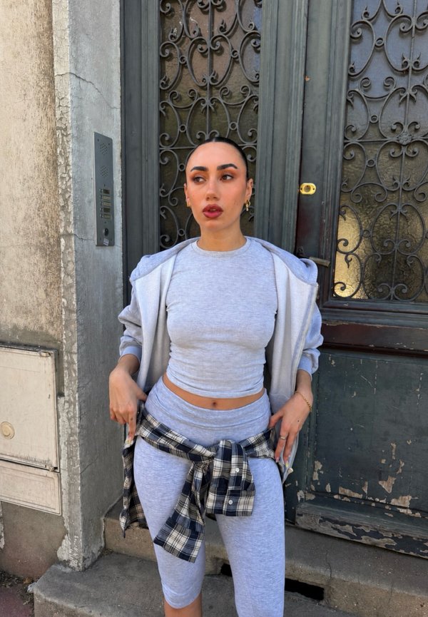 Donna che indossa un crop top grigio chiaro, leggings abbinati, una felpa con cappuccio grigia e una camicia a quadri annodata, in piedi davanti a una porta di metallo ornata.
