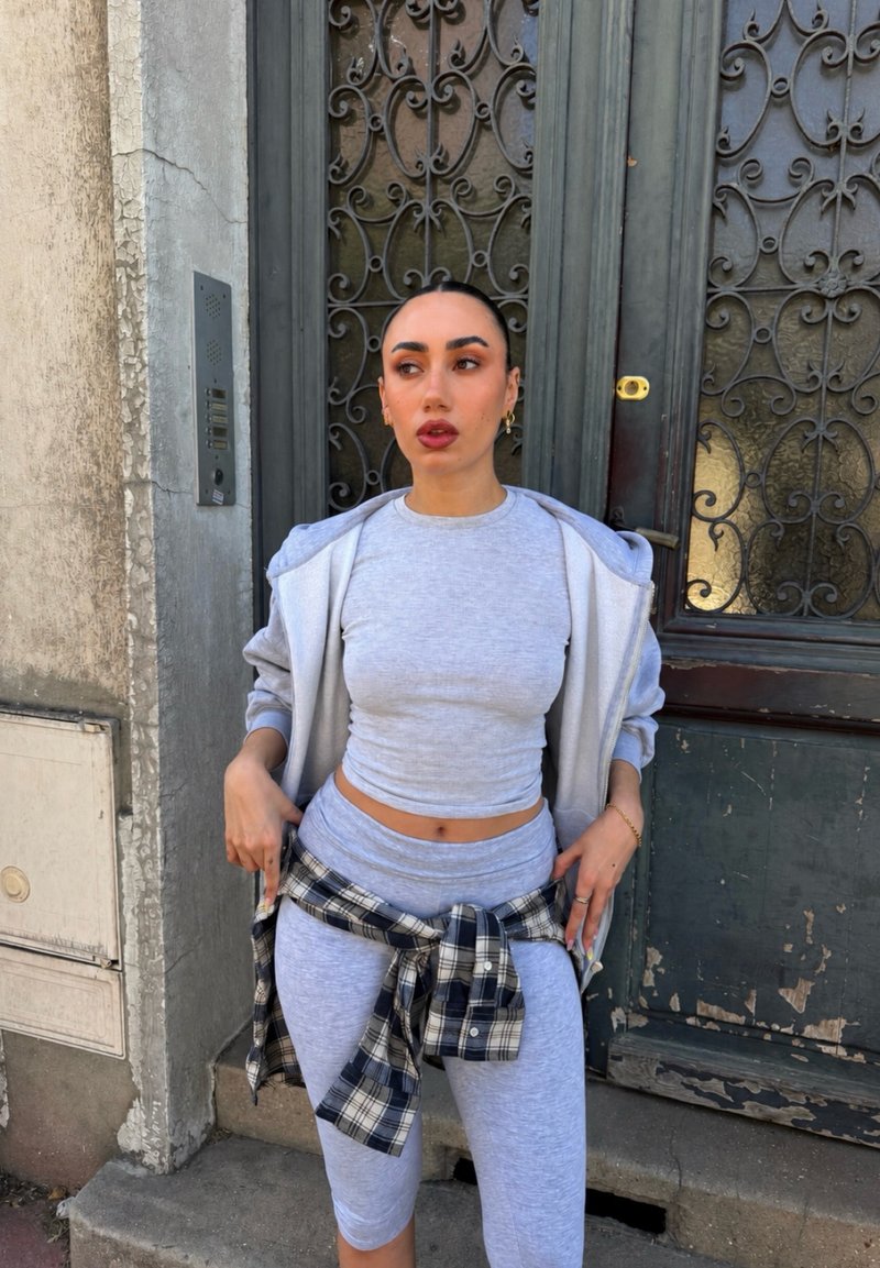 Mujer que lleva un top corto gris claro, leggings a juego, una sudadera con capucha gris y una camisa de cuadros anudada, de pie frente a una puerta metálica ornamentada.