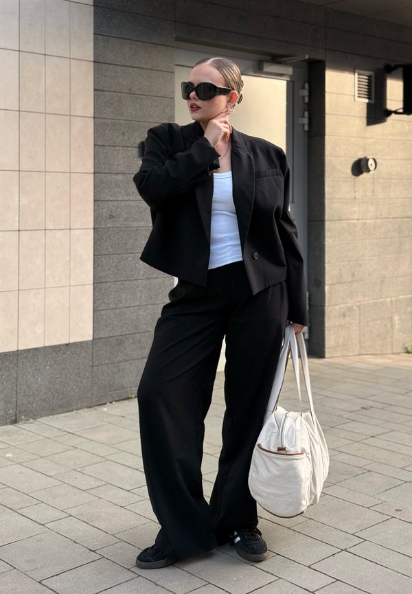 Femme en costume noir et haut blanc tenant un grand sac blanc, portant des lunettes de soleil noires, debout sur un trottoir carrelé près d'un bâtiment.