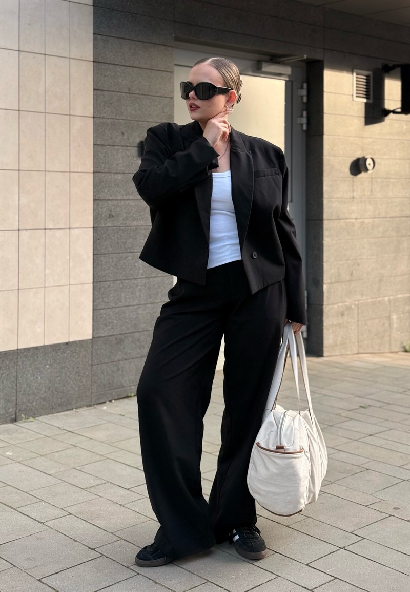 Femme en costume noir et haut blanc tenant un grand sac blanc, portant des lunettes de soleil noires, debout sur un trottoir carrelé près d'un bâtiment.