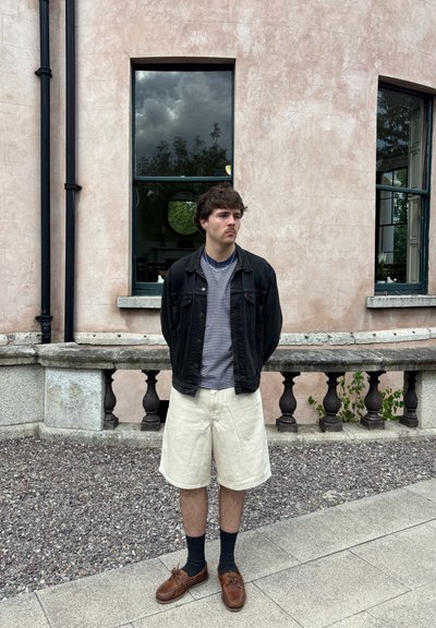 Chaqueta de mezclilla negra sobre una camiseta de rayas, pantalones cortos beige y zapatos marrones con calcetines oscuros. De pie contra una pared rosa texturizada con ventanas.