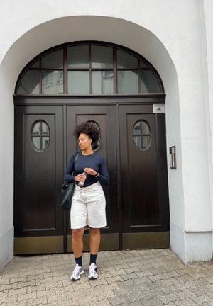 Puerta de madera negra con ventanas ovaladas, pantalones cortos blancos, camiseta de manga larga azul, bolso negro y zapatillas deportivas grises en un camino de adoquines.