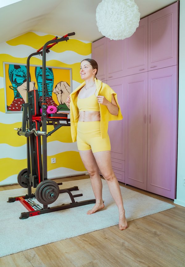 Femme en tenue de sport jaune avec une veste se prépare à faire de l'exercice à côté d'une machine de musculation dans une pièce colorée avec un mur à motifs et une armoire.