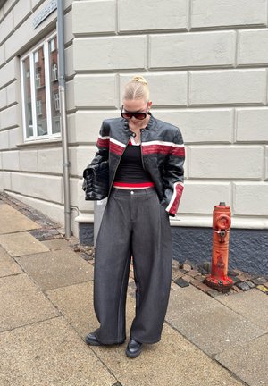 Mujer con chaqueta de cuero negra con rayas rojas y blancas, pantalones anchos grises, zapatos negros, gafas de sol y bolso negro, de pie en la acera.