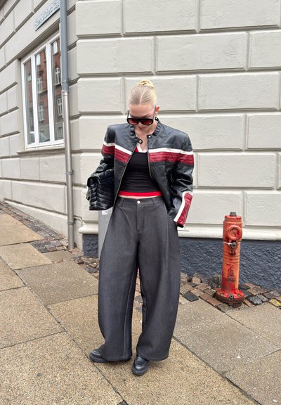 Mujer con chaqueta de cuero negra con rayas rojas y blancas, pantalones anchos grises, zapatos negros, gafas de sol y bolso negro, de pie en la acera.