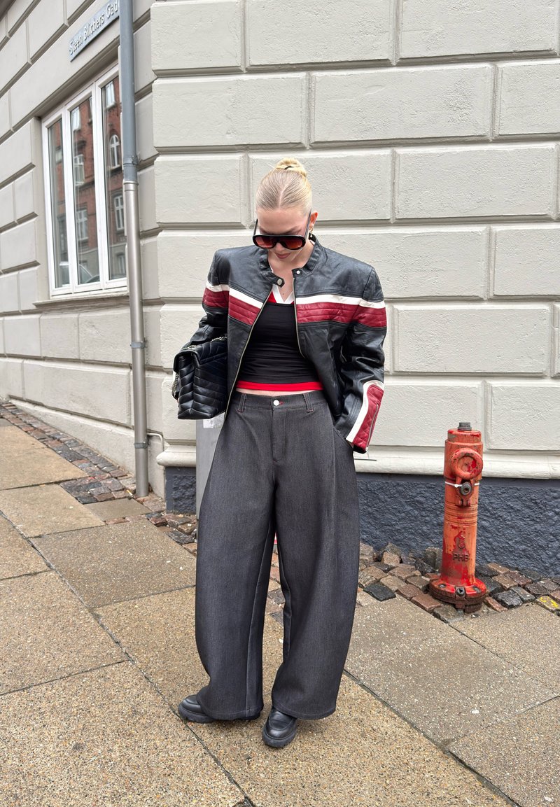 Femme en veste en cuir noire avec des rayures rouges et blanches, pantalon large gris, chaussures noires, lunettes de soleil et sac à main noir, debout sur le trottoir.