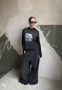 Čierna oversize košeľa s grafikou mačky, doplnená o široké sivé nohavice s textúrou. Modelka má na sebe čierne slnečné okuliare a vrstvový dlhý rukáv.