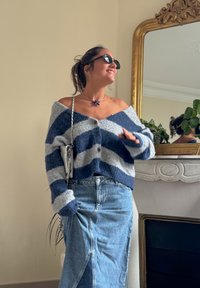 Debela pletena majica s mornarsko plavim i sivo prugama, s V-izrezom i detaljima na gumbima. Uparena s traper suknjom visokog struka. Ogledna pozadina.