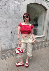 Top cropped vermelho com bolinhas brancas, calças cargo bege, mala da Hello Kitty em vermelho e branco, tamancos vermelhos com detalhes da Hello Kitty. Cabelo curto, óculos de sol.