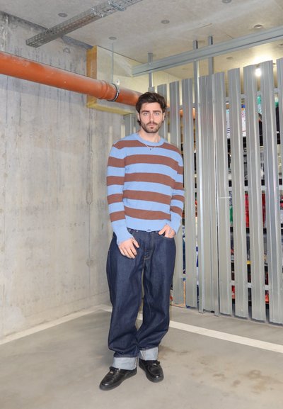Hombre con cabello oscuro y barba, vistiendo un suéter a rayas azul y marrón y jeans anchos remangados, de pie en una habitación industrial de concreto.