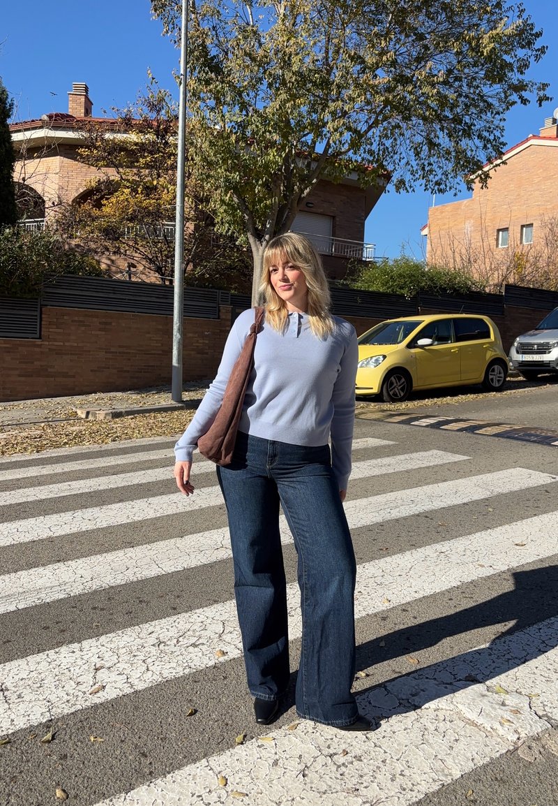 Femme aux cheveux blonds portant un pull bleu clair et un jean foncé, debout sur un passage pour piétons près de voitures stationnées et d'arbres par une journée ensoleillée.