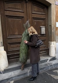Sapin enveloppé dans un filet, appuyé contre une porte en bois, avec une personne en manteau marron et bottes à talons noirs le touchant.