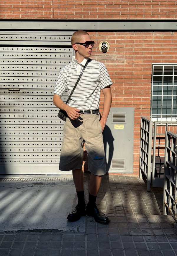 Junger Mann trägt gestreiftes Polohemd, beige Shorts, schwarze Socken und Schuhe, steht im Sonnenlicht vor einer Ziegel- und Metallwand.