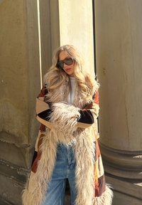 Lang, lappet frakk med krage av falsk pels i varme farger; lagd med en lett genser og høyhørende denimjeans; store solbriller.