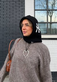 Femme portant une capuche en tricot noire avec un logo blanc, un pull marron duveteux, un pendentif en forme de cœur et un sac à bandoulière en cuir marron, debout près d'un mur en briques et d'une fenêtre.