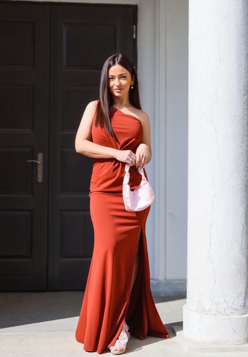 Woman in a sleeveless red dress with a thigh slit, holding a small pink handbag, standing near a white column and dark door.