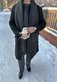 Personne portant un manteau foncé et une écharpe tenant deux gobelets de café à emporter et des gants noirs, debout sur un trottoir enneigé à côté d'une clôture en bois.