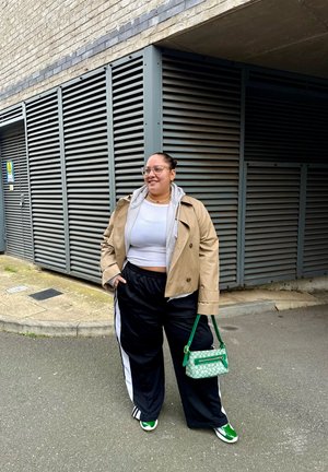Abrigo beige de doble botonadura sobre una sudadera con capucha gris, top corto blanco, pantalones deportivos negros con franjas blancas y zapatillas verdes. Bolso verde incluido.