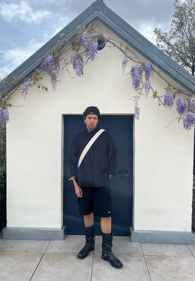 Persona vestida con una camisa negra de botones, pantalones cortos negros y botas altas negras hasta la rodilla, sostiene un bolso blanco cruzado y está de pie frente a un edificio blanco.