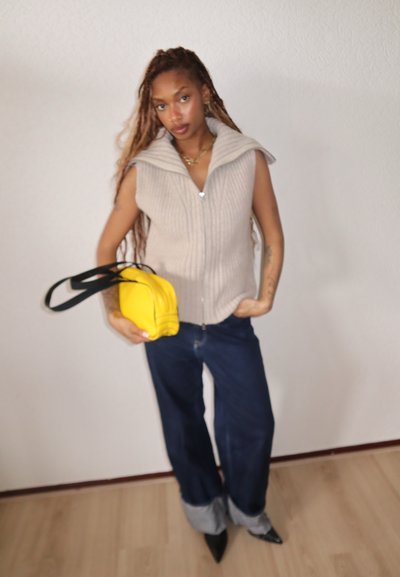 Mujer con trenzas largas que lleva un chaleco de punto sin mangas beige, vaqueros anchos de pierna con puños oscuros, zapatos negros puntiagudos y sostiene un bolso amarillo cruzado.