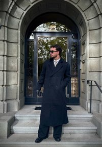 Man in a long black coat, gloves, and sunglasses stands on the steps outside a stone building featuring an arched doorway and a black-framed glass door.