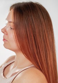 Jeune femme aux cheveux longs, raides et roux, portant un haut sans manches rose clair, regardant sur le côté sur un fond uni.