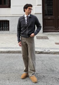 Veste zippée gris foncé avec col et poche, chemise blanche, cravate rayée, pantalon beige en velours côtelé et chaussures beiges à enfiler dans une rue.