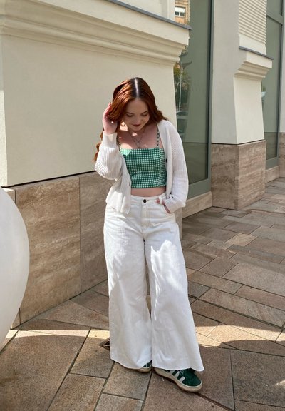 Pantalones anchos blancos combinados con un top corto a cuadros verdes y un cárdigan crema, contra un fondo de piedra clara.