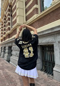 Maglia da football nera con "KAMARA" e numero "41" in oro. Gonna bianca a pieghe con un bordo giallo, in piedi contro un edificio in mattoni.