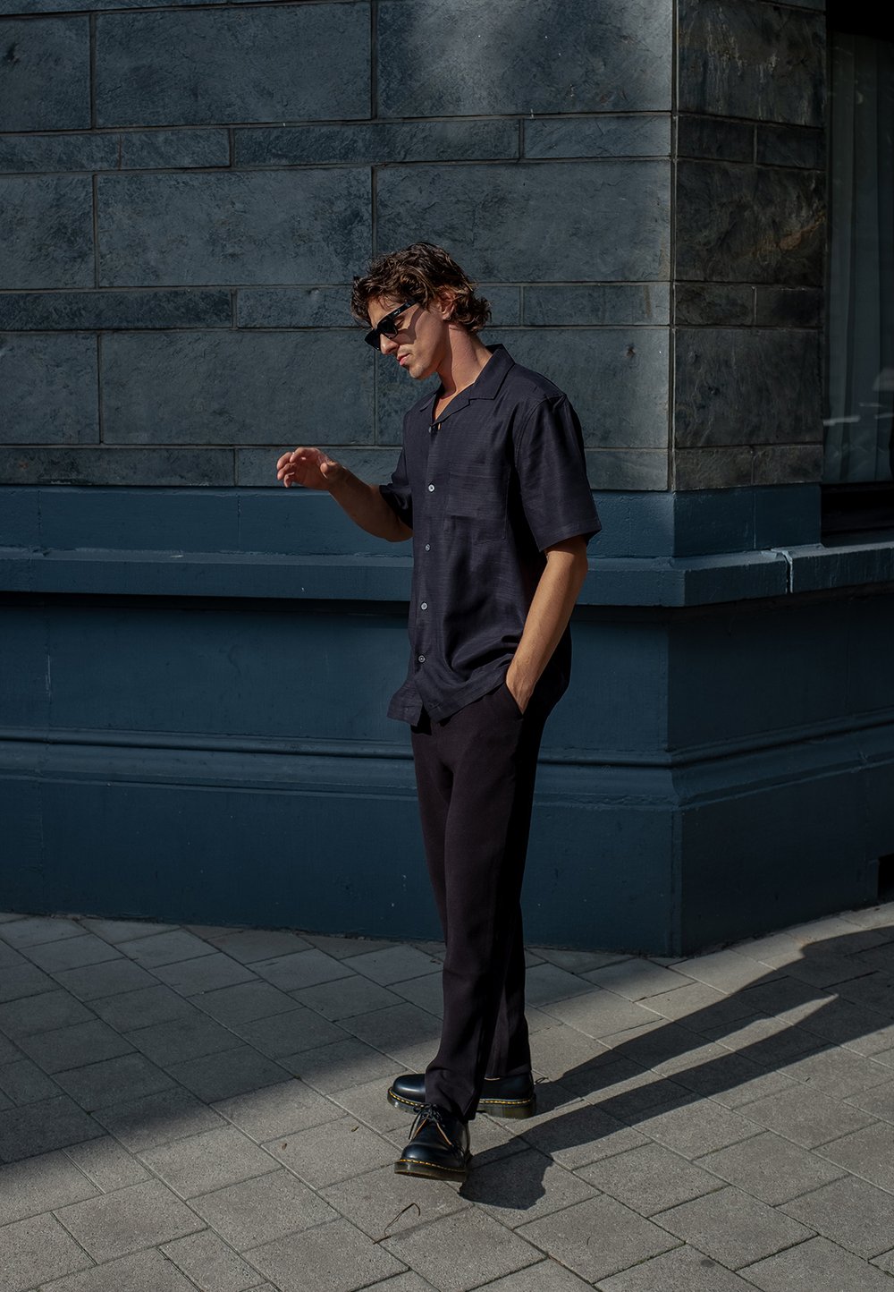 Svart kortärmad skjorta med en subtil textur, i kombination med mörka byxor och svarta skor. Modellen står mot en grå stenvägg.