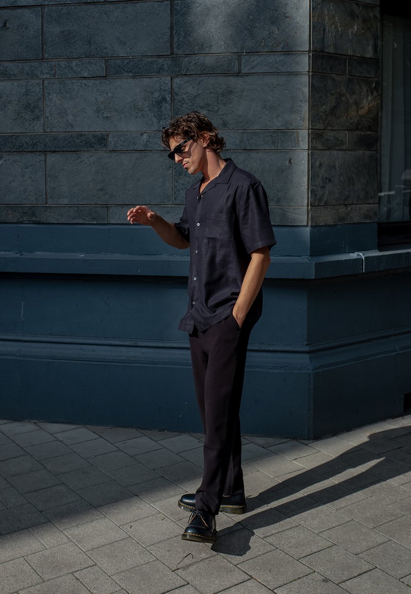 Juodas trumpas rankovės marškinėlis su subtiliu tekstūra, derinamas su tamsiais kelnais ir juodais batais. Modelis stovi prieš pilką akmeninę sieną.