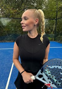 Černý těsný sportovní top, černá sukně, držící modrošedou pádla s perforacemi, stojící na modrém kurtu obklopená zelení.