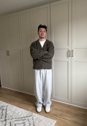 Cardigan marrón acanalado sobre una camiseta blanca, pantalones de chándal grises y zapatillas blancas. Está de pie frente a un armario de color claro.