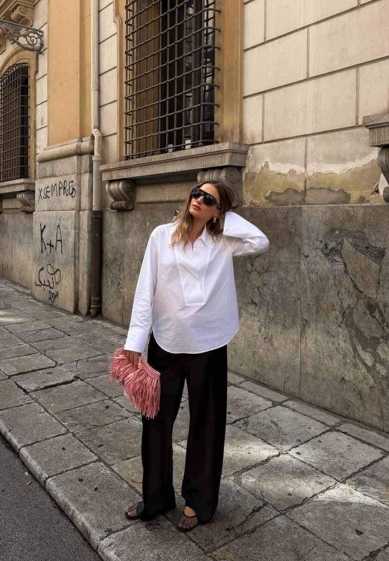Femme portant une chemise blanche et un pantalon noir, tenant un sac rose à franges, debout sur un trottoir en pierre à côté d'un bâtiment avec des graffitis.