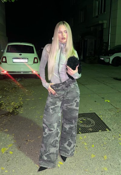 Pantalones anchos de camuflaje gris combinados con una blusa ajustada de cremallera gris claro, sostenida con un accesorio negro esponjoso, situados en una calle con poca iluminación.