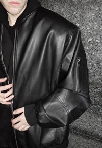 Jeune personne portant une veste en cuir noire par-dessus un sweat à capuche noir, les mains ajustant la fermeture éclair de la veste, debout devant un mur de pierre gris.