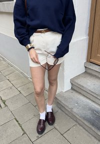 Marinblå sweatshirt, krämfärgade shorts, brun bälte med ett gult spänne, bruna loafers och vita ankelstrumpor. Håller bruna solglasögon.