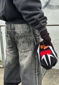 Felpa nera oversized, jeans grigi strappati con borchie metalliche e design simile a ali, che tiene un cappellino nero e rosso con dettagli bianchi.