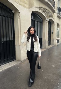 Hvid tekstureret cardigan med guldknapper, sort tætsiddende turtleneck, sorte flagrende bukser og en lille sort håndtaske. Urban indstilling med stenvægge.