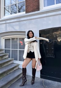Cardigan crema, top bianco con bottoni, gonna mini nera, stivali alti marroni con tacco. Tiene una piccola borsa rosa contro una porta scura.