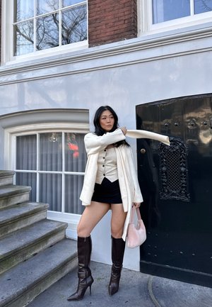 Cream cardigan, white top with buttons, black mini skirt, brown knee-high heeled boots. Holding a small pink bag against a dark door.