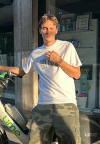Biela tričko s zeleným nápisom, maskáčové šortky, stojí vedľa Lime e-skutra so šedým a zeleným dizajnom, na jasnom slnku.