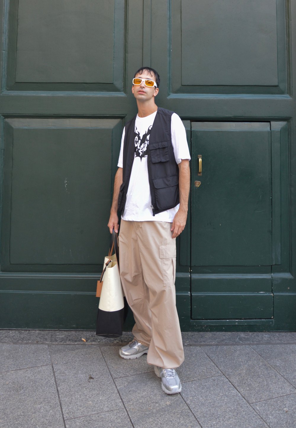 Chaleco negro sobre camiseta blanca con gráfico negro, pantalones cargo beige, zapatillas grises, sosteniendo una bolsa blanca y negra, contra un fondo verde.