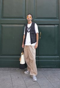 Chaleco negro sobre camiseta blanca con gráfico negro, pantalones cargo beige, zapatillas grises, sosteniendo una bolsa blanca y negra, contra un fondo verde.