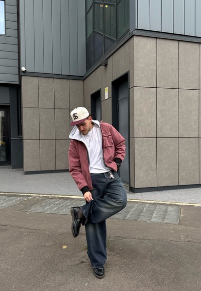 Chaqueta con capucha rosa sobre una camiseta blanca, Jeans anchos azul oscuro y zapatos negros. De pie frente a un edificio moderno con paneles grises.