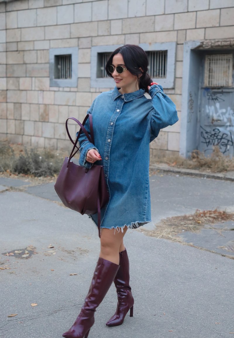 Denim blousejurk met kraag en knopen aan de voorkant, rafelige zoom, gecombineerd met kniehoge bordeauxrode laarzen en een bijpassende leren tas. Ronde zonnebril.