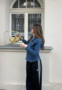 Denim jacket with buttons and pockets, black sports trousers with white stripes, holding a gold metallic handbag, standing by a window.